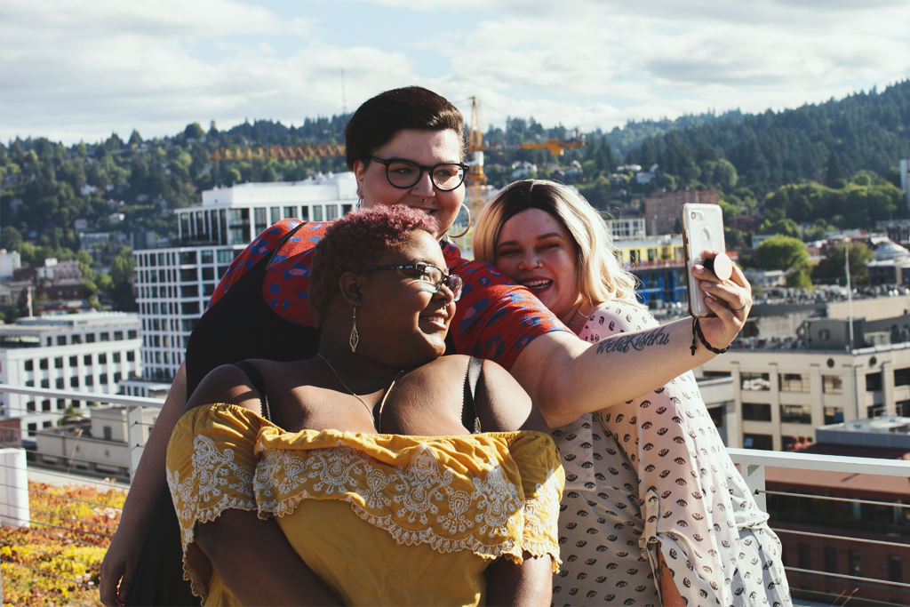 Framing fatness as a disease. Plus size women on rooftop with city view taking selfie with cell phone.