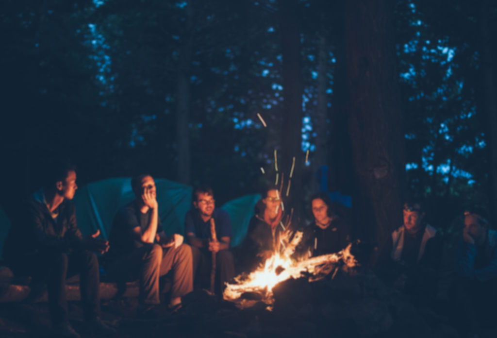 A group of people sitting by a campfire at night, listening to each other.