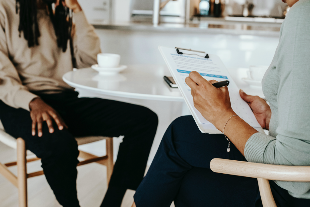 Differences between mental health professionals. A photo of a man and a woman sitting in front of each other. The woman is taking notes.