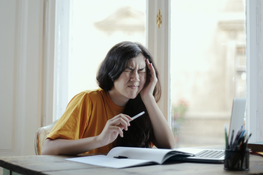 How to manage stress. A photo of a young woman sitting at a table with open books in front of her. She's holding a pen and has a hand on her head. Her eyes are closed and her face is scrunched up in an expression of pain.