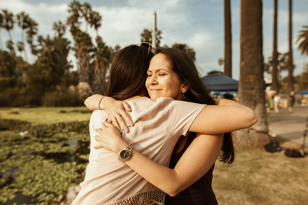 Suicide Prevention Awareness Day. A photo of two women hugging each other. They are standing outdoors in a park, you can see a lake and trees behind them.