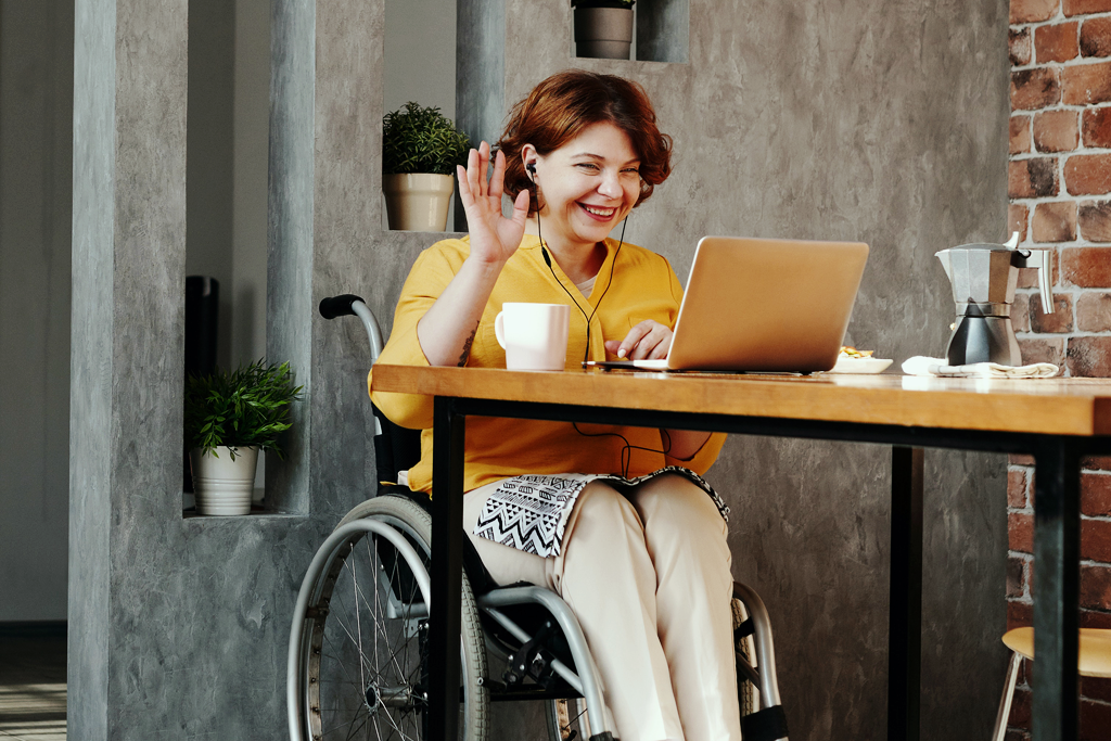 How well does telepsychology work? A photograph of a woman sitting on a wheelchair indoors in front of a computer. She's smiling and waving at the camera.