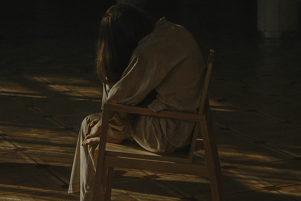 Impostor syndrome. A picture of a person with long hair, curled up in a chair in a dark room, facing away from the camera.