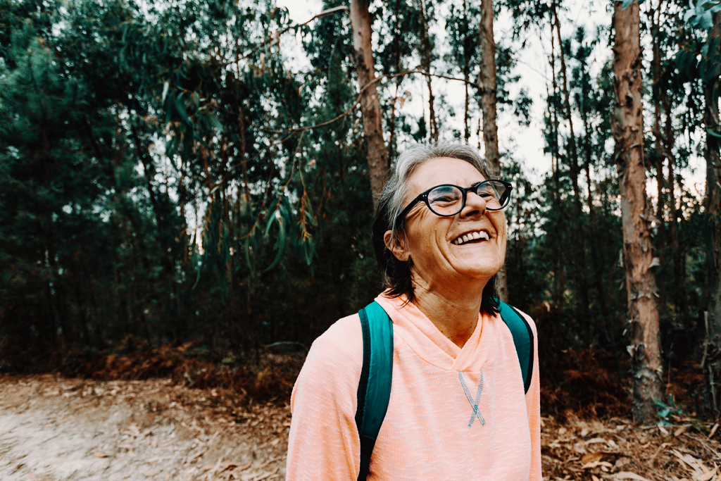 Hope as rebellion. Woman smiles wide and tilts her head back while standing outdoors with tall trees behind her.